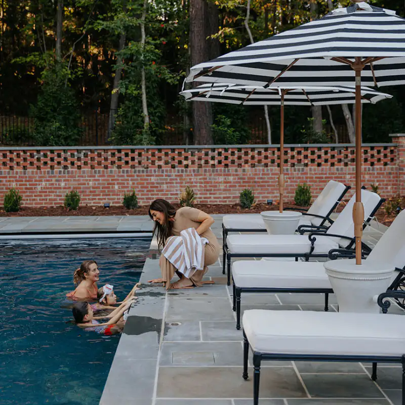 Friends relaxing on loungers beside a pool under a striped umbrella